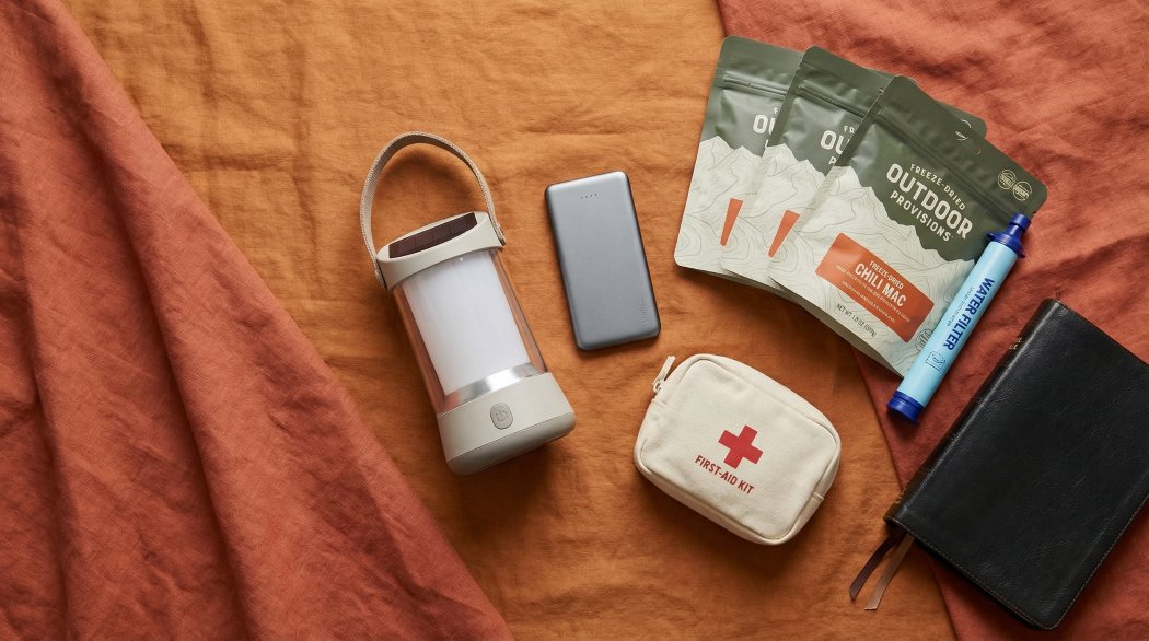 Modern flat-lay of emergency preparedness gear on an earth-tone background, including solar lantern, power bank, freeze-dried meals, water filter, first-aid kit, and a Bible.
